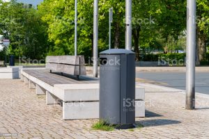 A metal trash bin and a long bench in an empty city park.